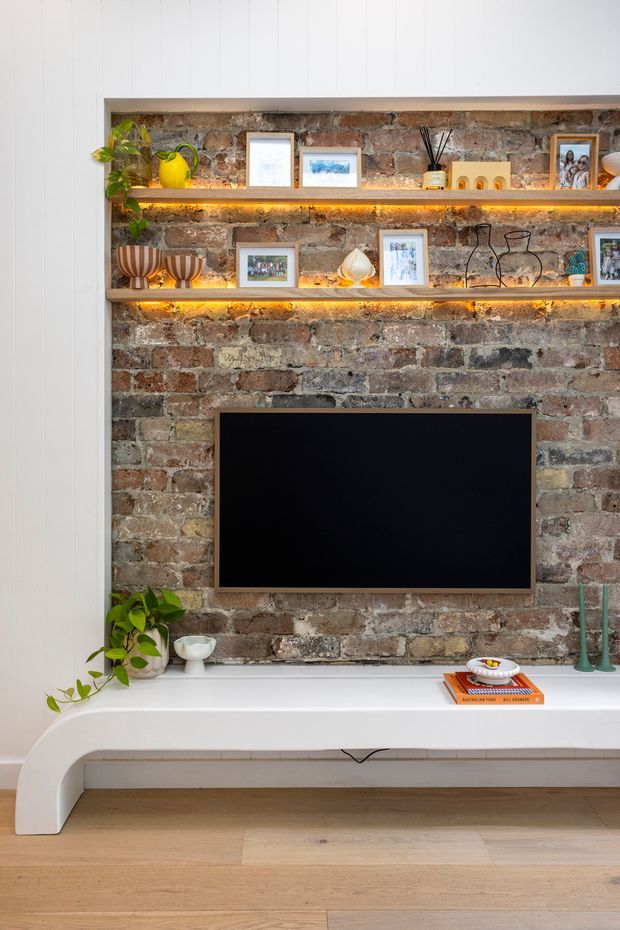 A styled living room media wall with an exposed red and grey brick feature wall, wall-mounted black television, illuminated timber display shelves, framed photographs and decorative objects. A long curved white built-in console runs beneath the television, with indoor plants and accessories, while pale timber flooring and white vertical wall panelling frame the composition.