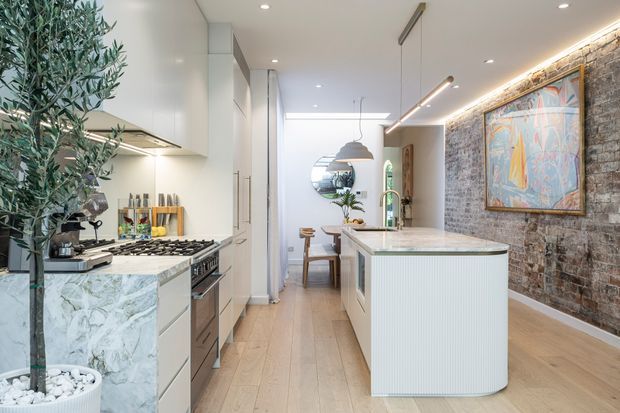 Bright, elongated contemporary kitchen with a central island, pale timber flooring, white integrated cabinetry, and a marble-look waterfall benchtop. A full-height exposed brick feature wall with framed artwork runs along the right side, while pendant and recessed lighting illuminate the kitchen and adjoining dining area. A large indoor olive tree and natural materials add warmth.