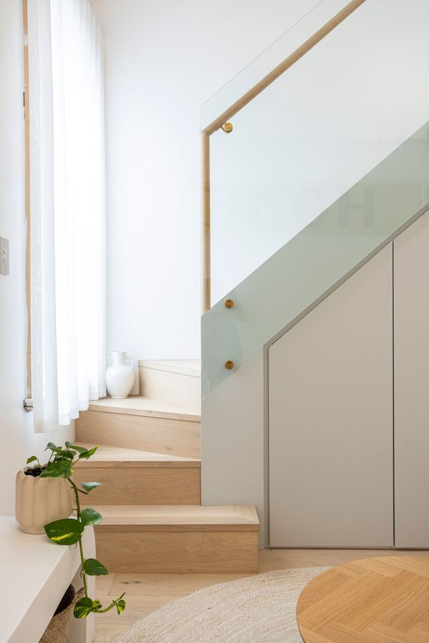 Bright, minimalist interior view of a compact stairway with pale timber stair treads and integrated cabinetry beneath the stairs. A clear glass balustrade is secured with warm brass-toned fittings, while sheer white curtains, white walls, a potted trailing plant, and a partial round timber table create a light, refined setting.