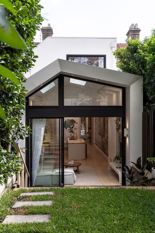 A contemporary rear extension viewed from a landscaped garden, combining a white upper volume with a sharply angled dark-framed glazed façade and projecting canopy. Large glass doors open into a bright interior with a sofa, kitchen island, timber flooring, and built-in shelving. Mature greenery frames the scene, while the original masonry chimney stacks and tiled roof remain visible behind the addition.