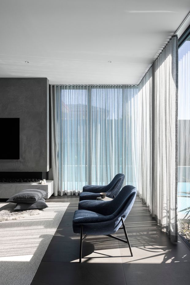 A refined contemporary living room with a dark stone or concrete feature wall and built-in television, two deep navy lounge chairs, a low coffee table, and a large pale area rug. Full-height sheer curtains line the corner glazing, creating strong filtered sunlight and shadows across the dark tiled floor. The space feels minimal, calm, and sophisticated.