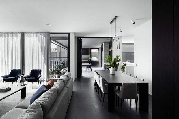 A refined open-plan contemporary interior combining a living room, dining area, and kitchen. The room features a large grey sectional sofa, dark dining table with upholstered chairs, black-framed internal glazing, sheer floor-to-ceiling curtains, dark tiled flooring, white walls and ceiling, and minimalist pendant and recessed lighting. A courtyard or glazed outdoor area is visible beyond the living space.