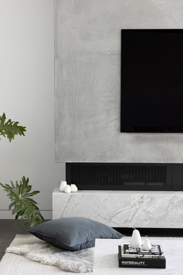 A refined contemporary living room vignette centred on a wall-mounted black television above a sculptural fireplace. The fireplace surround and mantel are clad in pale veined stone, with a textured light-grey plaster feature wall and a black patterned firebox or grille below. Soft grey cushions, a neutral textured rug, small white ceramic objects and leafy indoor plants create a calm, monochromatic setting.