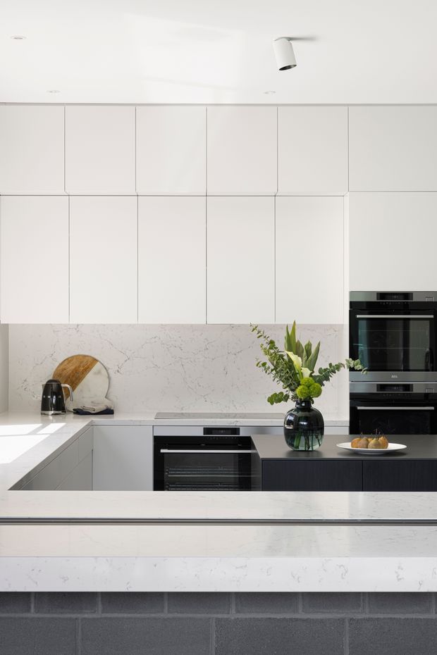 Bright minimalist kitchen with extensive white flat-panel cabinetry, a veined white stone backsplash and island benchtop, and integrated black appliances. A dark island base contrasts with the pale surfaces, while a vase of greenery and a small plate provide restrained styling. The kitchen is lit by recessed and white cylindrical ceiling lights.