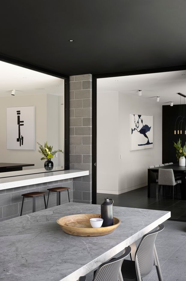 Contemporary open-plan kitchen and dining area with a large grey stone island, white waterfall-edge benchtop, dark cabinetry, grey blockwork feature wall, black-framed glazed partitions, and a black ceiling. Bar stools line the rear counter, while a dining table and pendant lighting are visible beyond the glazing. The space has a refined monochromatic palette with artwork and restrained floral arrangements.