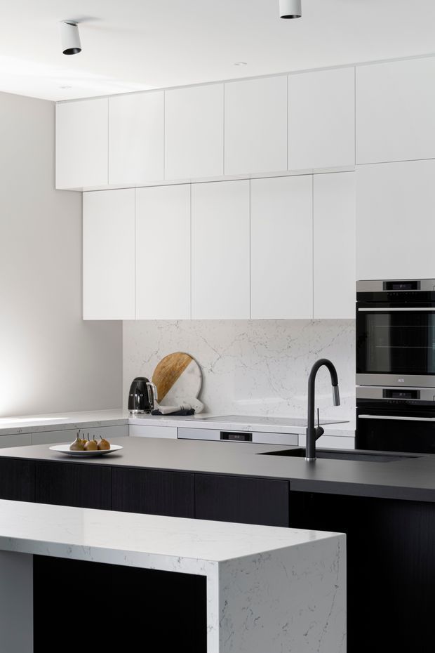 Bright, minimalist contemporary kitchen featuring extensive handleless white overhead cabinetry, dark timber-toned island cabinetry, pale marble-look benchtops and splashback, a matte black gooseneck tap, integrated dishwasher, and stacked black ovens. Recessed and cylindrical black ceiling lights add contrast to the otherwise restrained palette.