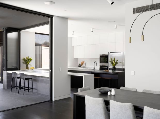 A bright, contemporary open-plan kitchen and dining area with a monochromatic black, white and grey palette. The kitchen features flat-panel white overhead cabinetry, dark lower cabinetry, a dark island benchtop, integrated appliances and a minimalist black tap. A large dark dining table with upholstered grey chairs occupies the foreground. Black-framed sliding glass doors open to a covered outdoor entertaining area with a raised bar and stools. Curved black pendant arms with brass-toned fittings add a sculptural detail above the dining area.