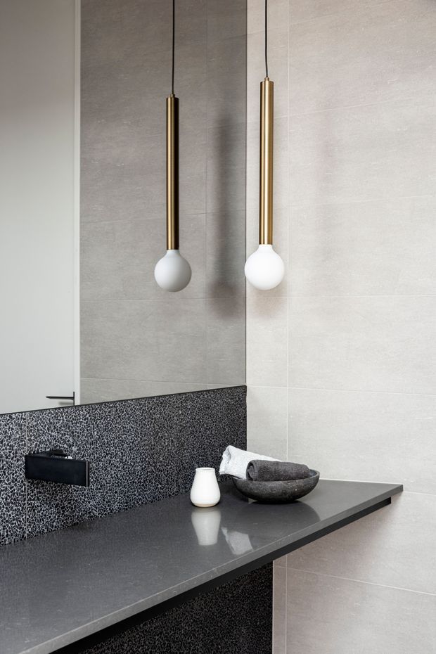 Close-up view of a refined contemporary bathroom vanity. Large pale grey wall tiles surround a dark charcoal benchtop and matching dark textured mosaic splashback, with a black wall-mounted tap fitting, vessel bowl, and two slim brass-and-white pendant lights reflected in the mirror. The restrained grey, black, white, and brass palette creates a polished, minimalist atmosphere.