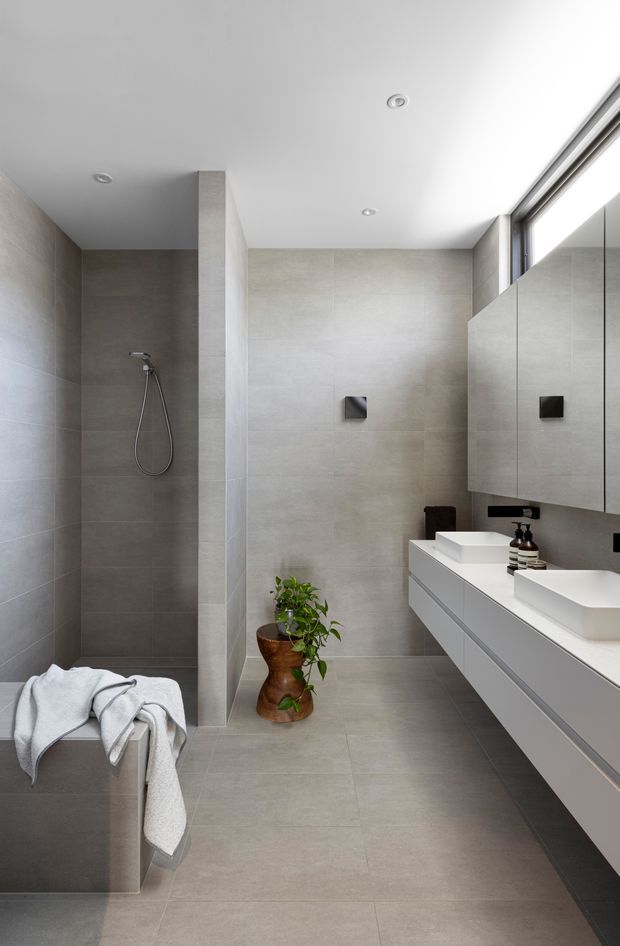 A contemporary bathroom with a walk-in shower, large-format grey-beige tiled walls and floor, a floating double vanity with white integrated basins, mirrored cabinetry, recessed ceiling lights, and a high horizontal window. A small potted plant and folded towels add natural and soft accents.