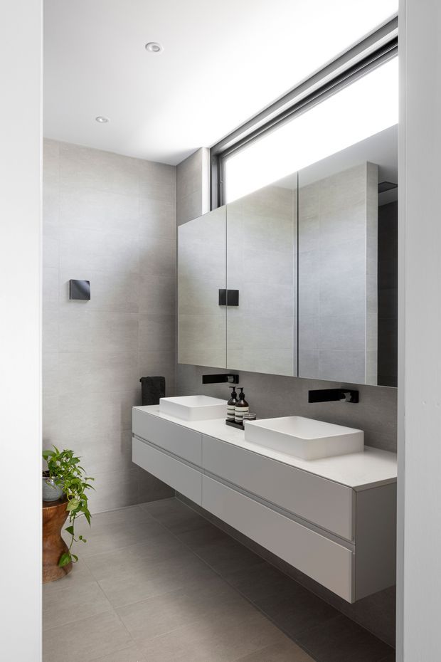 A bright, contemporary double-vanity bathroom with a floating light-grey wall-mounted vanity, two white vessel basins, black wall-mounted tapware, and mirrored cabinetry above. Large-format pale grey tiles cover the walls and floor, while a high horizontal window brings in natural light. Potted greenery adds a small natural accent.