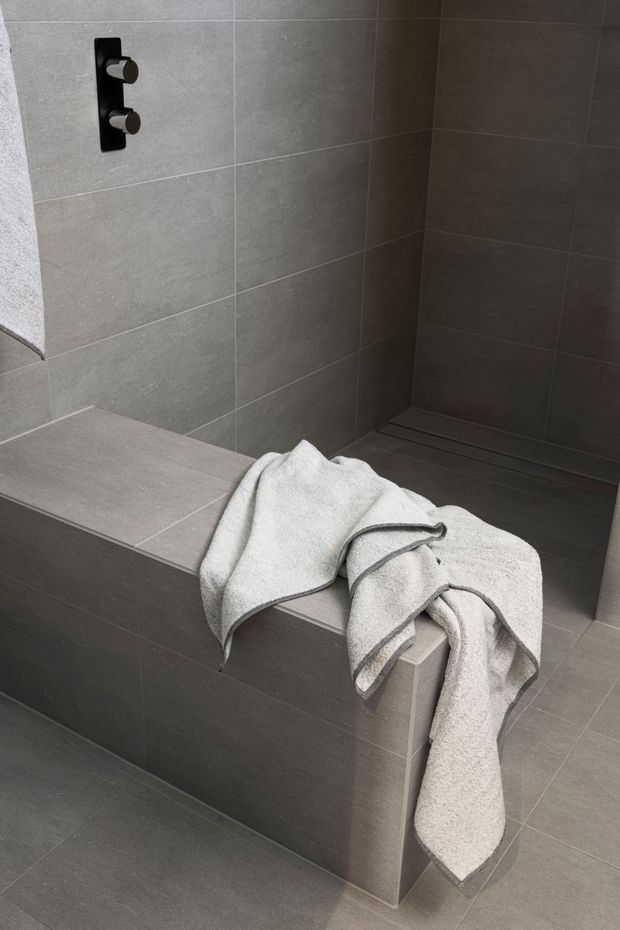 A minimalist walk-in shower with a built-in tiled bench and dark concealed shower controls. Large-format grey tiles cover the walls, bench, floor, and shower platform, creating a monochromatic, refined bathroom interior. A light grey towel is draped over the bench in the foreground.