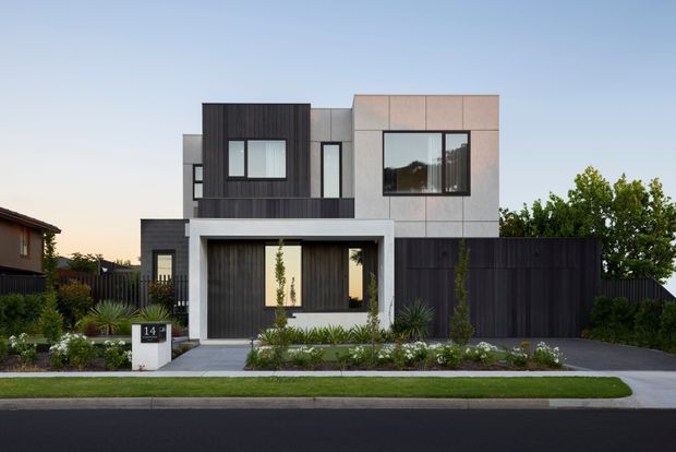 A contemporary multi-storey urban residence viewed from the street, with a strongly geometric facade composed of dark vertical cladding, pale grey rendered or panelled volumes, and large black-framed windows. The front entry is recessed beneath a projecting upper-level element, with a dark garage or side enclosure, landscaped garden beds, a paved path, and a neat suburban streetscape.
