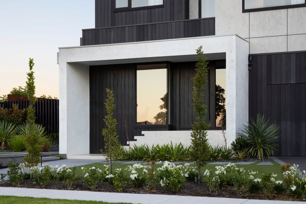 Contemporary multi-storey residential facade with a strongly geometric composition. The exterior combines dark vertical cladding, pale rendered or concrete-look wall planes, large black-framed windows, and a projecting entry canopy. A landscaped front garden with ornamental grasses, shrubs, and low flowering plants sits beside a paved path and dark vertical boundary fencing.