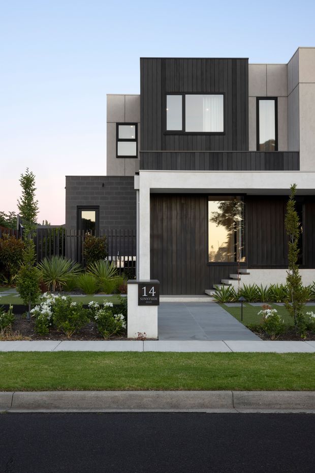 Front exterior view of a contemporary multi-storey townhouse with a strong geometric composition. The facade combines dark vertical cladding, light grey rendered or panelled surfaces, black brickwork, large rectangular windows and a recessed entry. A neat landscaped front garden with ornamental grasses, shrubs, paving and lawn leads to the front door, with a numbered address plaque beside the path.