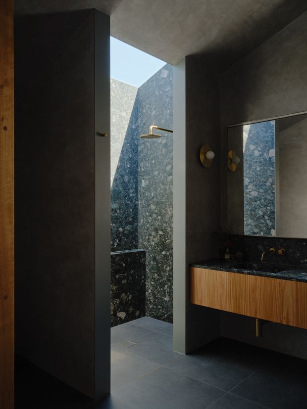A dark, sophisticated contemporary bathroom featuring a walk-in shower open to an overhead skylight or lightwell. The shower walls use dramatic blue-grey natural stone with prominent aggregate veining, while the room has dark plastered walls, large dark floor tiles, a timber floating vanity with a dark stone benchtop, wall-mounted brass-toned lighting, and a large mirror.