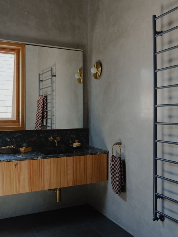 A moody, refined bathroom vanity area with a floating vertically grained timber cabinet, dark stone benchtop and splashback, wall-mounted brass tapware, large frameless mirror, timber-framed window, and a dark metal heated towel rail. Textured grey plaster walls and dark floor tiles create a warm, dramatic atmosphere, with brass wall lighting and accessories providing accents.
