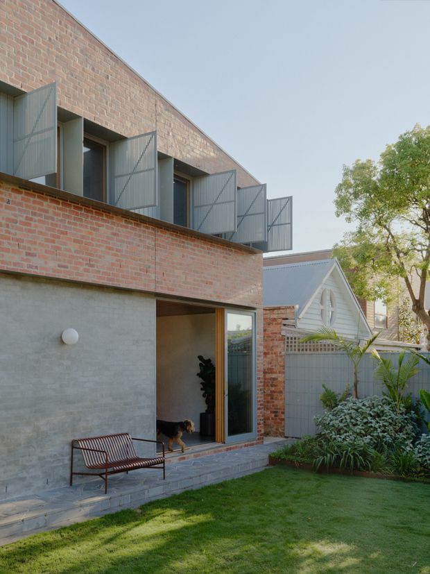 Exterior view of a contemporary two-storey brick residence opening onto a landscaped courtyard garden. The facade combines exposed red brick with a smooth grey concrete lower level, timber-framed glazing, and projecting upper-level windows screened by grey perforated metal shutters. A small paved terrace with a metal bench leads to the glazed doorway, while a neighbouring gabled structure and established planting are visible beyond.