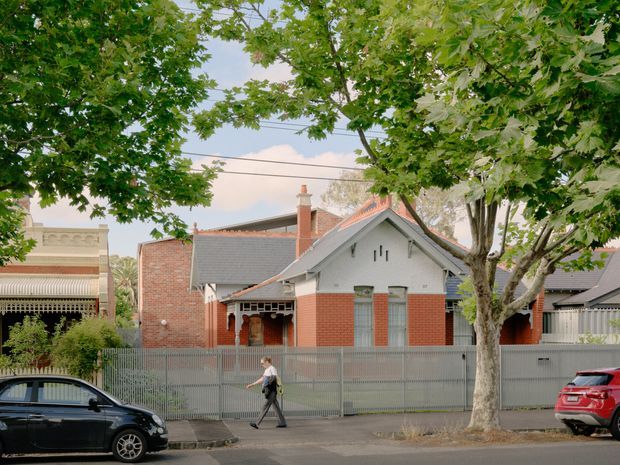 Street-facing view of a charming single-storey heritage cottage, with red brick walls, grey-painted gabled frontage, decorative eaves, small vertical windows, and a covered entry porch behind a metal front fence. Mature street trees frame the house, with neighbouring period buildings and parked cars visible along the suburban road.