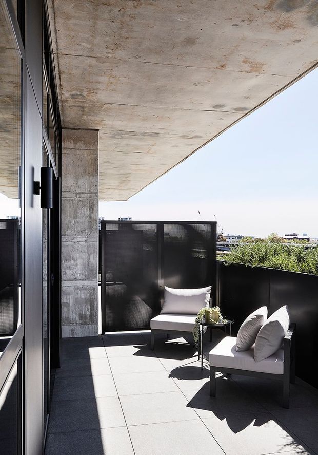 A modern elevated outdoor terrace or balcony with dark-framed glazing, exposed raw concrete soffit and column, large grey floor tiles, black privacy screens and a low planter. Two light upholstered lounge chairs and a small side table create a compact seating area, with bright daylight and distant urban views beyond.
