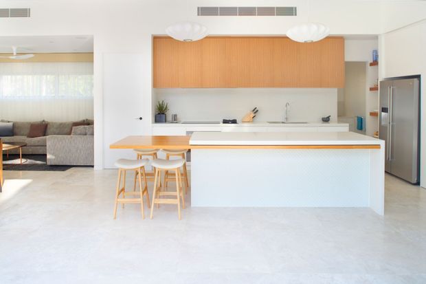 Bright open-plan kitchen with a large white island, integrated timber breakfast bar and matching timber stools. The kitchen features flat-panel warm timber overhead cabinetry, white benchtops, a pale textured island front, stainless steel refrigerator, and polished light flooring. A living room is visible through the opening to the left.