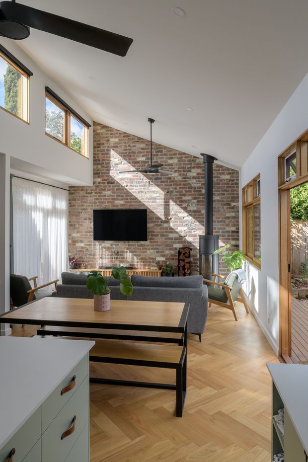 Bright open-plan living and dining area viewed from the kitchen, with a high sloping white ceiling, exposed red-brick feature wall, large timber-framed windows and doors, herringbone timber flooring, grey sofa, timber dining table, black wood-burning stove and ceiling fans. Sunlight creates strong geometric shadows across the brick wall, and a deck and greenery are visible through the glazing.
