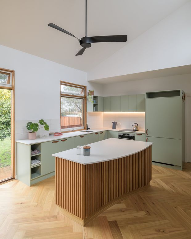 Bright contemporary kitchen with a large curved island featuring vertical timber fluting, pale green built-in cabinetry, integrated appliances, white tiled splashback, and warm herringbone timber flooring. Tall timber-framed windows bring in abundant natural light, while a black ceiling fan provides a strong overhead feature.