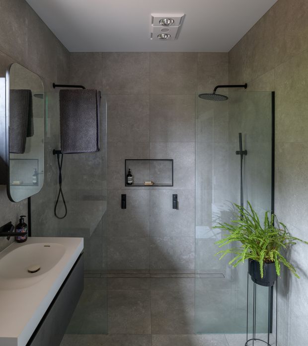 Contemporary bathroom with large-format grey stone-look wall and floor tiles, a frameless glass walk-in shower, wall-mounted black shower fittings, recessed niche shelving, a floating dark vanity with white basin, rounded mirror, and a potted fern.