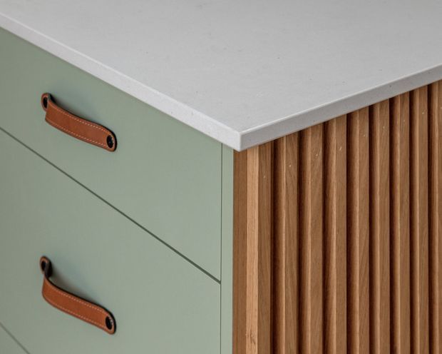 Close-up detail of built-in cabinetry showing muted sage-green drawer fronts with brown leather pull handles, a white stone-like benchtop, and a vertical natural timber slatted feature panel. The image focuses on fixed joinery and material detailing.