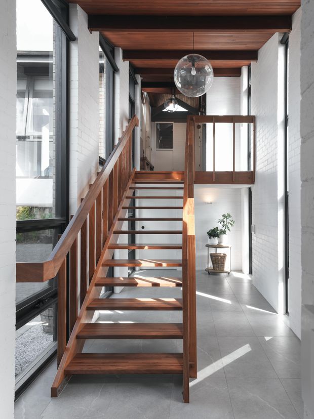 A bright, modern interior stairway with open-riser timber stairs and matching timber balustrades. White-painted brick walls, large black-framed windows, grey tiled flooring, exposed dark timber ceiling beams and slatted timber ceiling panels create a strong architectural rhythm. Clear glass globe pendant lights hang above the stair and landing, while a potted plant sits in the naturally lit hallway below.