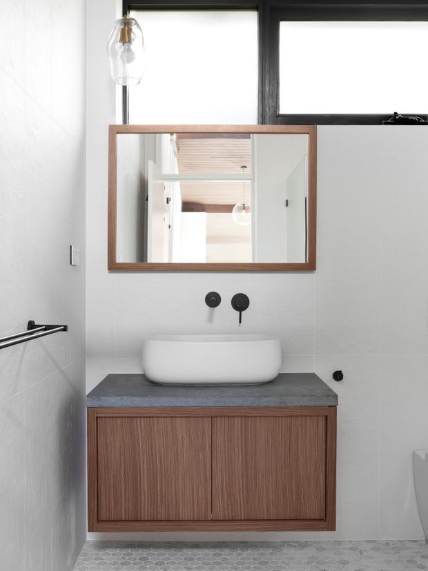 Compact contemporary bathroom vanity area with a white vessel basin on a dark grey stone benchtop, warm timber-grain wall-mounted cabinetry, a matching timber-framed mirror, white tiled walls, and small grey hexagonal floor tiles. Black tapware, window frames, and hardware provide contrast, while a glass pendant light hangs above.