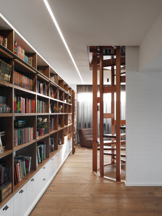 A bright, narrow interior passage dominated by a full-height built-in timber bookcase with white lower cabinetry and open shelving filled with books. A sculptural timber stair or balustrade structure occupies the centre-right, leading toward a partially visible living area with a sofa and sheer curtains. The space has pale timber flooring, white walls and ceiling, and a linear recessed ceiling light.