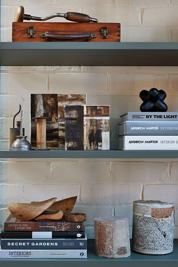 Close-up interior vignette of dark grey built-in shelving set against a white-painted textured brick wall. The shelves display stacked design books, abstract art panels, a vintage hand drill, a small metal vessel, wooden sculptural objects, and cylindrical concrete or terrazzo-like vessels. The styling is curated, industrial and earthy, with strong tactile materials and restrained neutral tones.