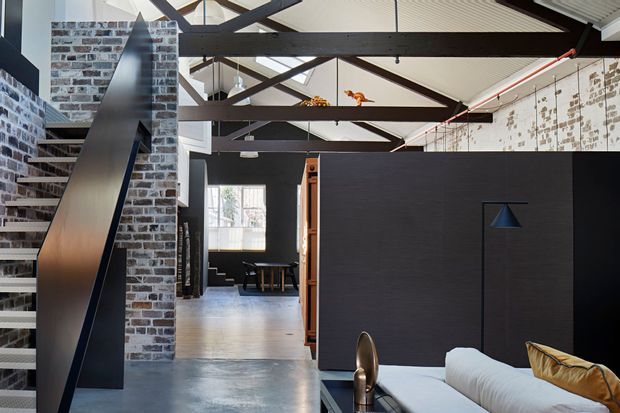 Open-plan industrial-style interior with a high, exposed-beam ceiling. A dark steel stair and balustrade rises beside exposed brick walls, while polished concrete flooring, black feature walls, and visible ductwork reinforce the warehouse-like character. A living area with a pale sofa is visible in the foreground, with dining or workspace elements beyond.