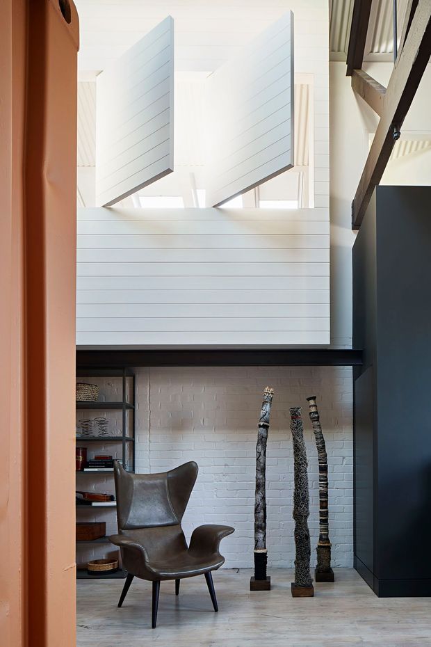 A bright, double-height interior featuring white horizontal wall panelling, a white-painted brick fireplace wall with a dark timber mantel, and high clerestory-style openings with angled projecting panels. A sculptural leather lounge chair sits beside open shelving, while tall decorative objects stand near a large dark built-in joinery element. Pale timber flooring and strong natural light create a refined, airy contemporary setting.