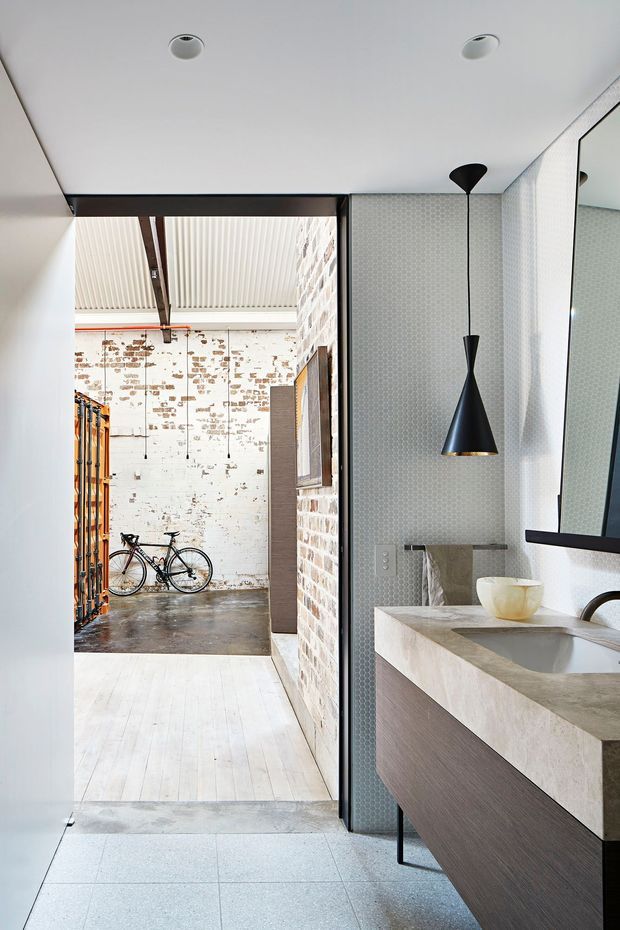 A contemporary bathroom vanity occupies the right side of the image, with a large concrete or stone benchtop, integrated basin, wood-toned floating cabinetry, black-framed mirror, and a sculptural black pendant light. Pale patterned wall tiles and light floor tiles create a refined neutral interior. Through the open doorway is an industrial-style adjoining space with exposed brick walls, concrete flooring, exposed roof structure, and a bicycle.