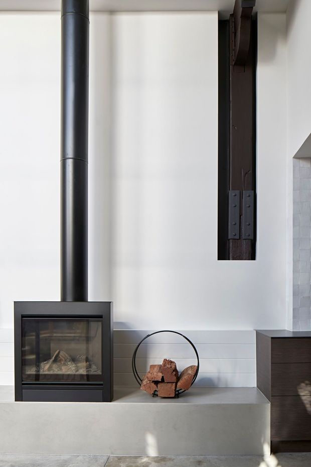 A bright minimalist interior focused on a contemporary black wood-burning fireplace with a tall cylindrical flue. The fireplace sits on a smooth raised concrete hearth, beside a circular black metal firewood holder. White horizontal wall panelling, a dark exposed timber structural element, and partial dark built-in cabinetry are visible.