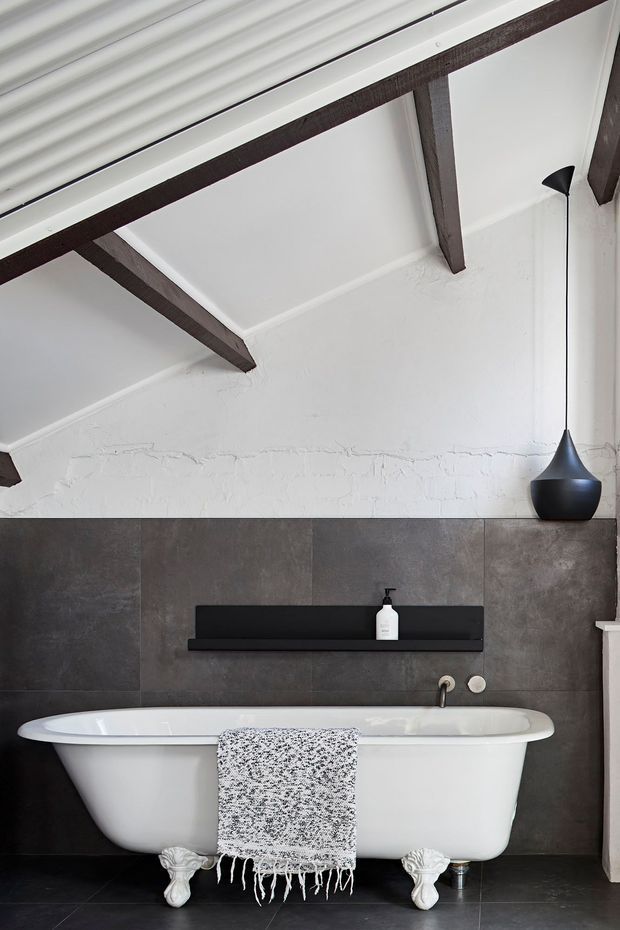 A refined bathroom with a freestanding white clawfoot bathtub positioned against a dark charcoal tiled feature wall. The room has white plaster walls, exposed dark timber ceiling beams, a black wall-mounted shelf, minimalist black pendant-style lighting, and dark floor tiles. A textured monochrome throw is draped over the bath, creating a dramatic yet elegant contrast.