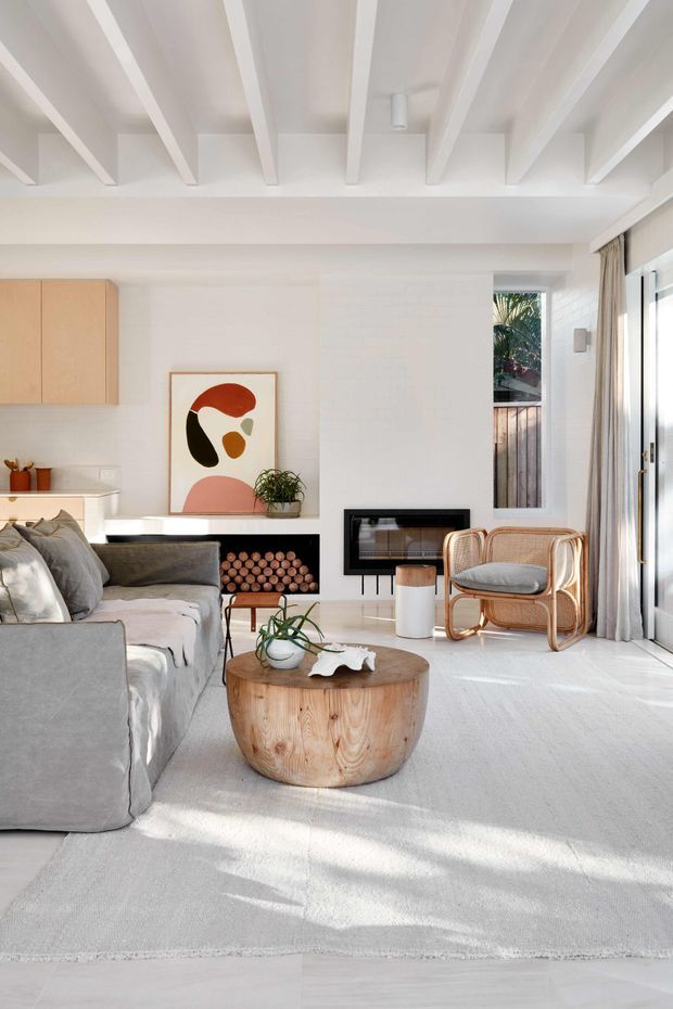 Bright contemporary living room with a white-painted exposed-beam ceiling, white brick-style feature wall, integrated black fireplace, pale built-in cabinetry, large glazed doors with curtains, a grey sofa, curved timber coffee table, and rattan lounge chairs. The room has abundant natural light and a soft neutral palette with small earthy artwork accents.