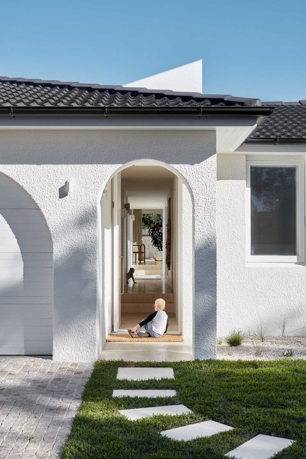 Bright exterior view of a white rendered residential house with a dark tiled pitched roof, guttering, arched entry portal, and a glazed hallway visible through the open doorway. A paved driveway, stepping-stone path, and lawn lead to the entrance. The facade has a clean Mediterranean-influenced form with strong sunlight and deep shadows.