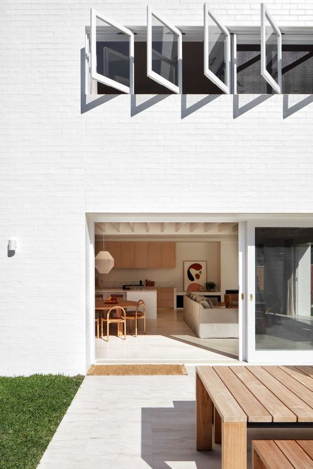 Bright exterior view of a contemporary white-painted brick facade with a row of projecting awning windows above large glazed sliding doors. The open doors reveal a warm, light-toned open-plan kitchen and living area, while a timber outdoor dining table and deck occupy the foreground. Strong sunlight creates crisp geometric shadows across the facade and paving.