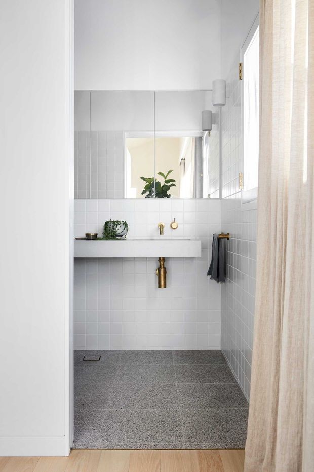 A bright, compact contemporary bathroom viewed from a timber-floored hallway or adjacent room. The room features white square wall tiles, a grey terrazzo-like tiled floor, a floating white vanity with an integrated basin, wall-mounted brass tapware and exposed brass waste pipe, a large frameless mirror, cylindrical wall lights, a narrow window, and a light neutral curtain. A small plant and dark towel add restrained accents.