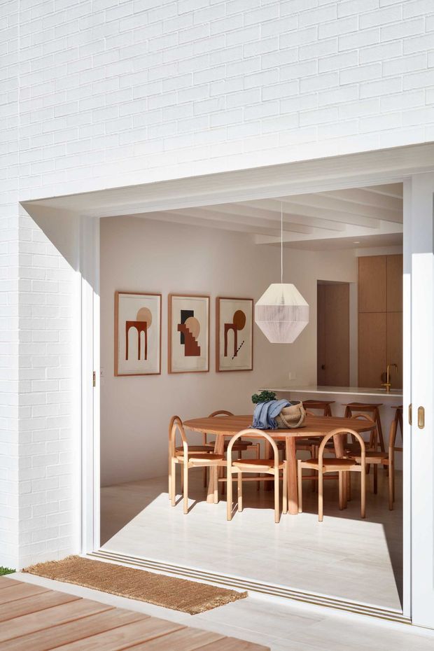 A bright dining area viewed from an exterior timber deck through wide open sliding doors. The room features a light timber dining table and curved-back chairs, a faceted white pendant light, framed abstract artwork, pale walls and ceiling, and timber built-in cabinetry with a visible kitchen counter beyond. White painted brick surrounds the opening, with strong natural sunlight creating a warm, airy atmosphere.