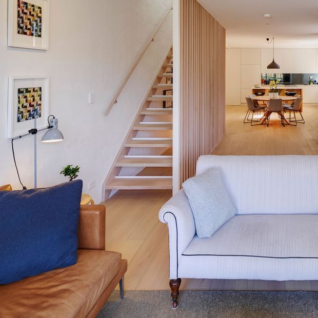 Bright open-plan residential interior showing a living room in the foreground, a timber staircase, a vertical timber slat divider, and a dining and kitchen area beyond. The room features pale timber flooring, a light upholstered sofa, a tan leather chair, blue cushions, white walls, framed artwork, and simple contemporary furnishings.