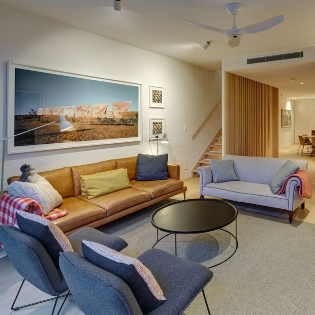 A spacious, light-filled living room with a warm mid-century character. The room features a tan leather sofa, pale blue upholstered sofa and armchairs, a low round black coffee table, framed wall artwork, and a large area rug. A timber staircase and vertical timber screen lead toward a dining area, while a ceiling fan and recessed air-conditioning provide functional details.