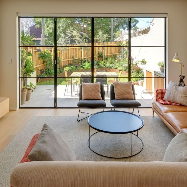 Bright, contemporary living room viewed toward a landscaped courtyard through full-width black-framed sliding glass doors. The room contains a large neutral sofa, two mid-century-style lounge chairs, a round black coffee table, a floor lamp, and a pale textured rug. Outside is a paved patio with a dining table, timber fencing, and dense planting. The interior has light timber flooring, warm off-white walls, and abundant natural light.