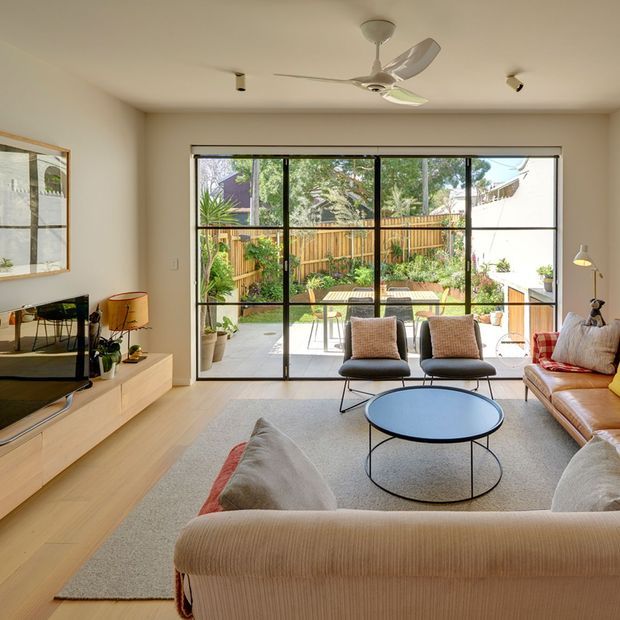 A bright, contemporary living room viewed from behind a large sofa, with a low round coffee table, two lounge chairs, a leather sofa, media console and television. Full-height black-framed glazing opens the room visually to a small planted courtyard and paved outdoor area. Pale timber flooring, light neutral walls, a ceiling fan and warm natural light create an airy, relaxed interior.