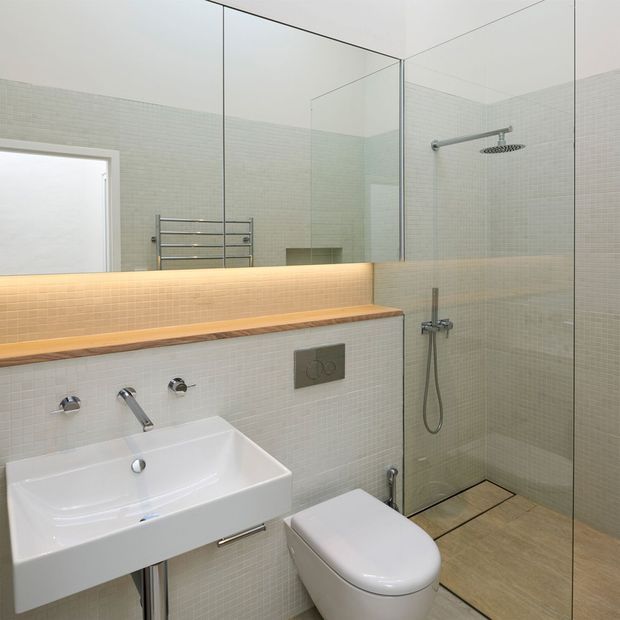 Contemporary bathroom with a wall-mounted white basin, wall-hung toilet, large frameless glass shower enclosure, chrome fittings, heated towel rail, and pale small-format tiled walls. A warm timber ledge and softly illuminated tiled vanity backsplash add contrast to the otherwise light, minimalist space.