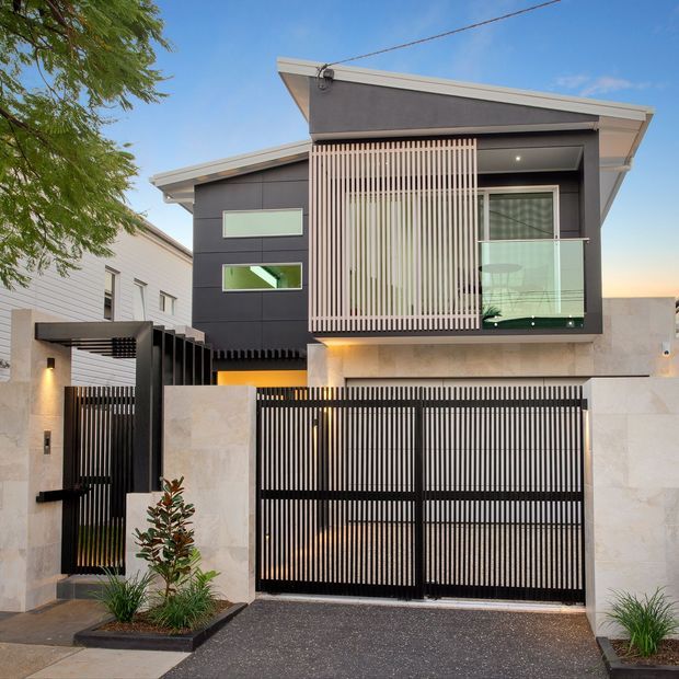 Street-facing modern two-storey townhouse facade with dark cladding, a low-pitched skillion-style roof, large glazing, vertical timber-look screening, a glass balcony balustrade, and a black slatted garage and pedestrian gate. Pale stone boundary walls, charcoal paving, and restrained landscaping frame the entry.