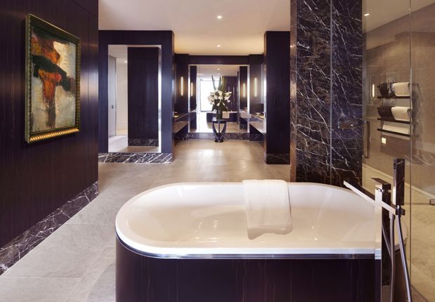 Luxurious contemporary bathroom featuring a large freestanding oval bathtub, dark timber wall panelling, dramatic dark marble accents, pale stone flooring, and a glass-enclosed shower with open shelving. A corridor and vanity area are visible beyond the main bathing space, with warm recessed lighting and refined metallic fixtures.