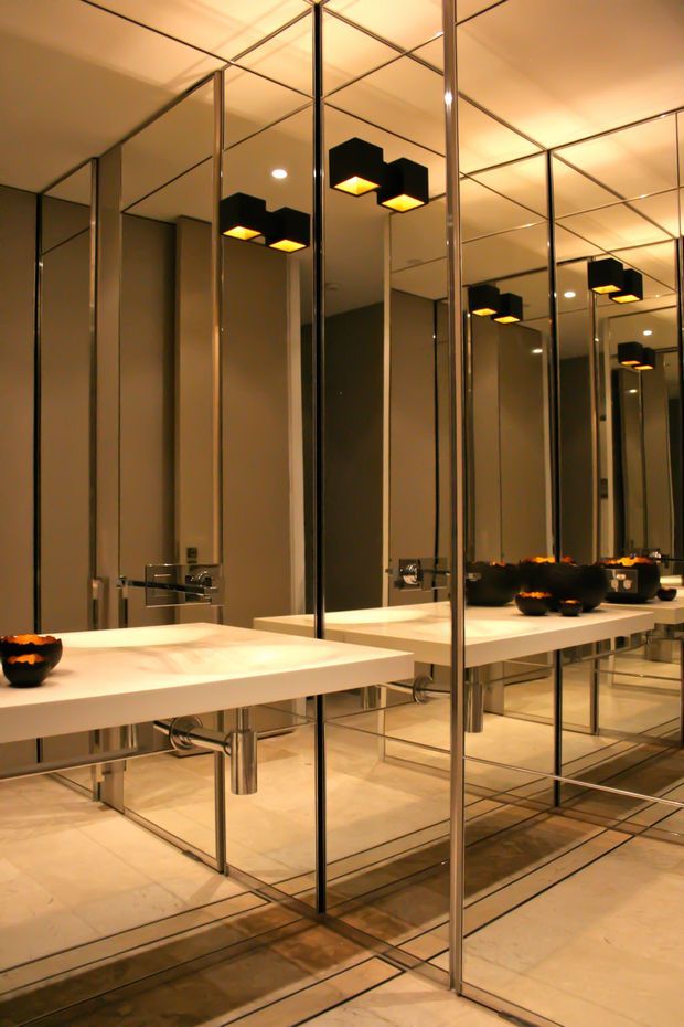 A dramatic contemporary bathroom or powder room with floor-to-ceiling mirrored wall panels, a long white floating vanity, wall-mounted chrome faucets, and under-mounted basins. Warm recessed and geometric black pendant lighting is reflected repeatedly in the mirrors, creating a highly reflective, luxurious atmosphere. Light stone or porcelain floor tiles and dark decorative bowls accent the space.